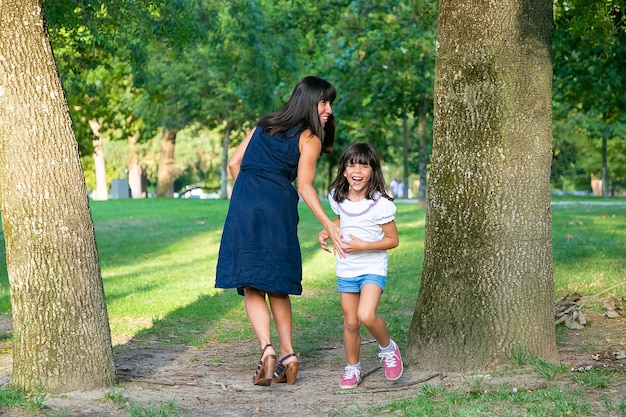 Niña feliz emocionada jugando juegos activos con su mamá al aire libre, de pie junto a los árboles en el parque y riendo. Longitud total. Concepto de ocio y actividad familiar al aire libre