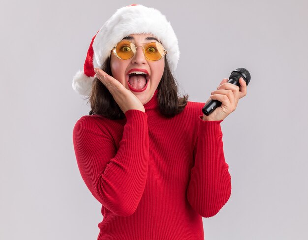Niña feliz y emocionada en suéter rojo y gorro de Papá Noel con gafas sosteniendo micrófono mirando a la cámara de pie sobre fondo blanco.