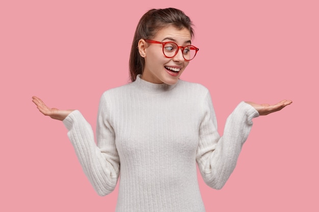 Niña feliz extiende las manos, finge sostener algo, mira con alegría a un lado, vestida con un jersey blanco, gafas de borde rojo