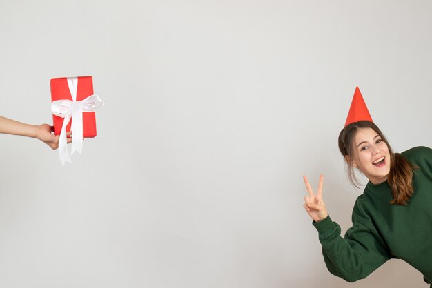 Niña feliz con gorro de fiesta haciendo regalo de signo de victoria en mano humana sobre blanco