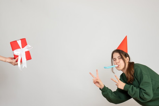 Niña feliz con gorro de fiesta haciendo el signo de la victoria con matraca mano humana sosteniendo el regalo en blanco