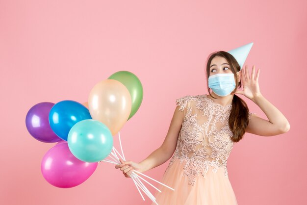 niña feliz con gorro de fiesta y máscara médica escuchando algo mientras sostiene globos de colores en rosa