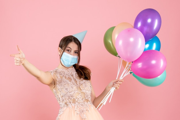 niña feliz con gorro de fiesta y máscara médica sosteniendo globos de colores en rosa