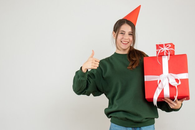 Niña feliz con gorro de fiesta y regalos haciendo pulgar arriba firmar en blanco