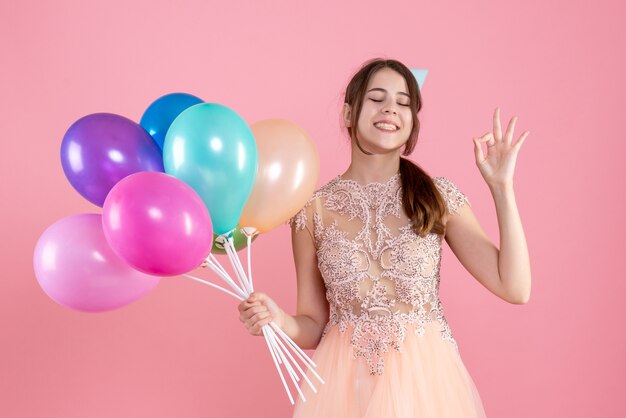 niña feliz con gorro de fiesta sosteniendo globos cerrando los ojos en rosa