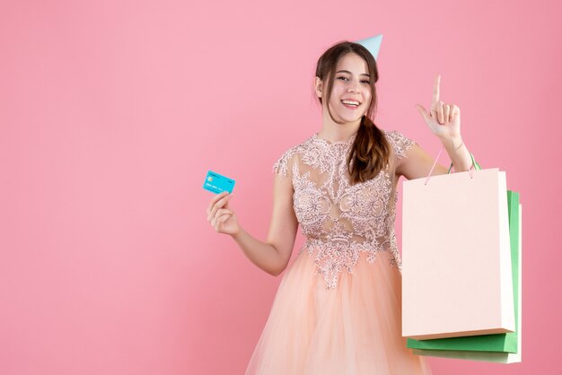 niña feliz con gorro de fiesta con tarjeta y bolsas de compras en rosa