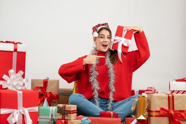 Niña feliz con gorro de Papá Noel apuntando al regalo sentado alrededor presenta en blanco