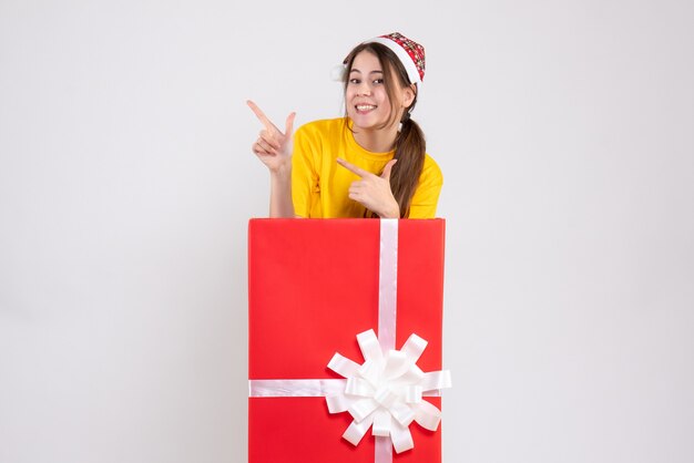 Niña feliz con gorro de Papá Noel apuntando a algo detrás de un gran regalo de Navidad en blanco