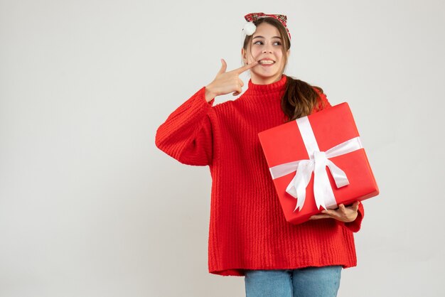 Niña feliz con gorro de Papá Noel apuntando a su sonrisa sosteniendo un regalo en blanco