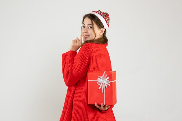 Niña feliz con gorro de Papá Noel escondiendo regalo de Navidad detrás de su espalda en blanco