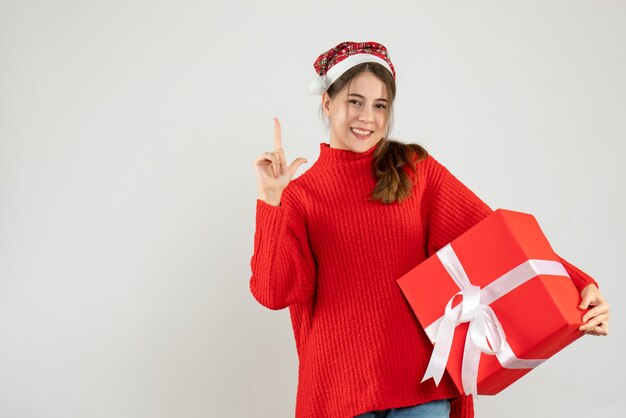 Niña feliz con gorro de Papá Noel haciendo pistola de dedo sosteniendo regalo en blanco