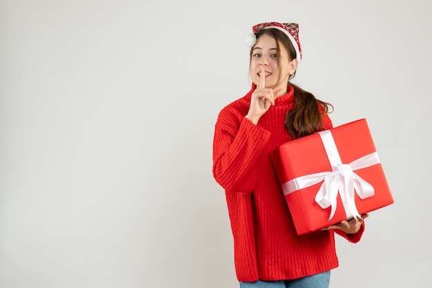 niña feliz con gorro de Papá Noel haciendo shh firmar con regalo en blanco