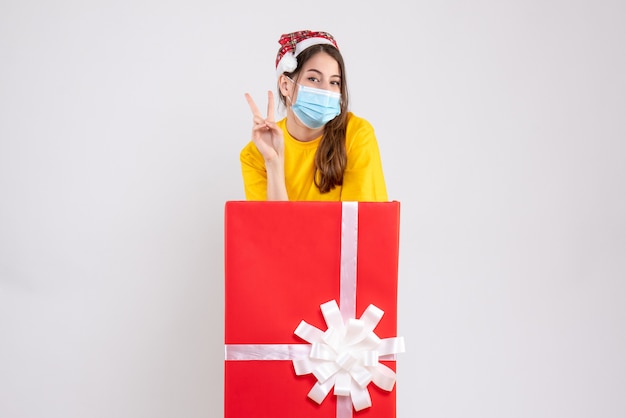 Niña feliz con gorro de Papá Noel haciendo el signo de la victoria de pie detrás de un gran regalo de Navidad en blanco