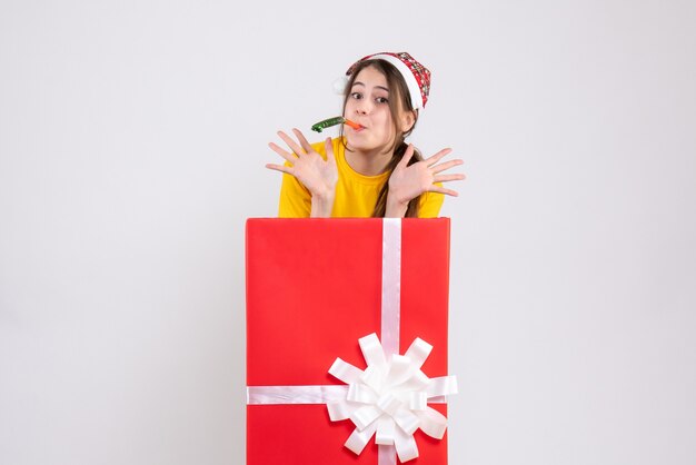 Niña feliz con gorro de Papá Noel con matraca de pie detrás de un gran regalo de Navidad en blanco