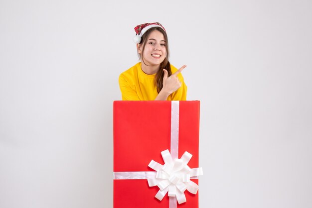 Niña feliz con gorro de Papá Noel mostrando algo de pie detrás de un gran regalo de Navidad en blanco