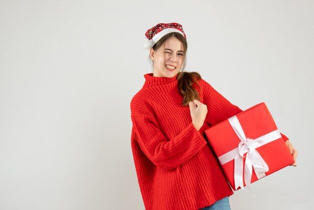 Niña feliz con gorro de Papá Noel mostrando gesto ganador sosteniendo regalo en blanco