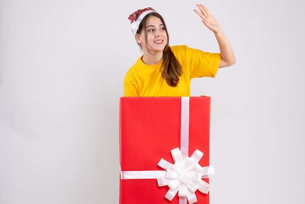 Niña feliz con gorro de Papá Noel de pie detrás de un gran regalo de Navidad en blanco