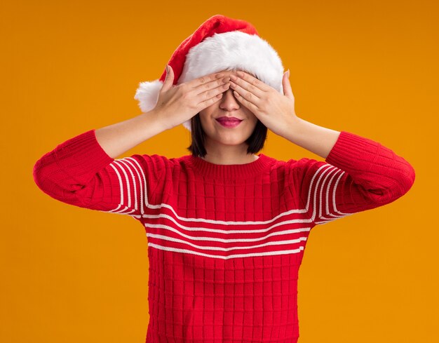 Niña feliz con gorro de Papá Noel que cubre los ojos con las manos aisladas en la pared naranja