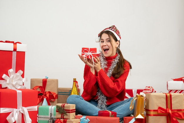 Niña feliz con gorro de Papá Noel sosteniendo presente con ambas manos sentados alrededor de regalos en blanco