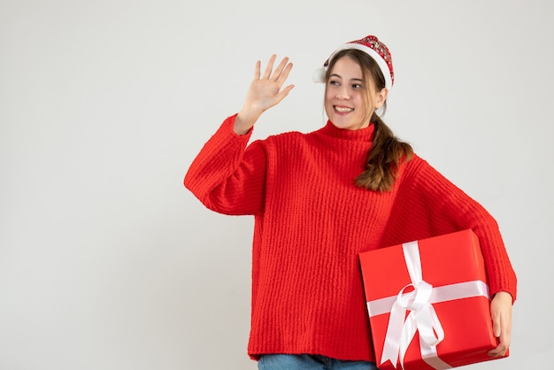 niña feliz con gorro de Papá Noel sosteniendo un regalo y saludando a alguien en blanco