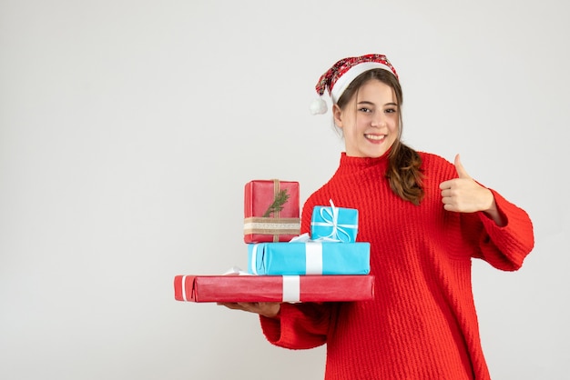 Niña feliz con gorro de Papá Noel sosteniendo su regalo de Navidad haciendo gesto con el pulgar hacia arriba en blanco