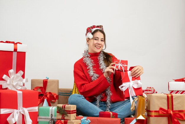 Niña feliz con gorro de Papá Noel sosteniendo sus regalos sentados alrededor de regalos en blanco