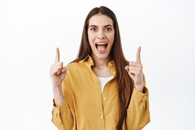 Niña feliz grita y muestra una oferta de promoción increíble en la parte superior, apuntando con el dedo hacia arriba y sonriendo ampliamente, demostrando algo genial, de pie sobre una pared blanca