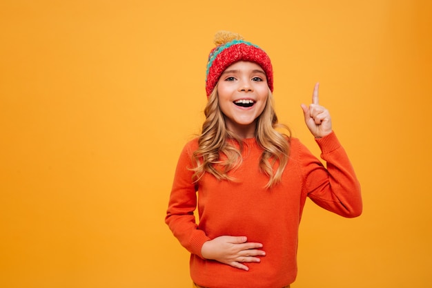 Niña feliz hambrienta en suéter y sombrero sosteniendo su barriga y teniendo idea mientras mira a la cámara sobre naranja