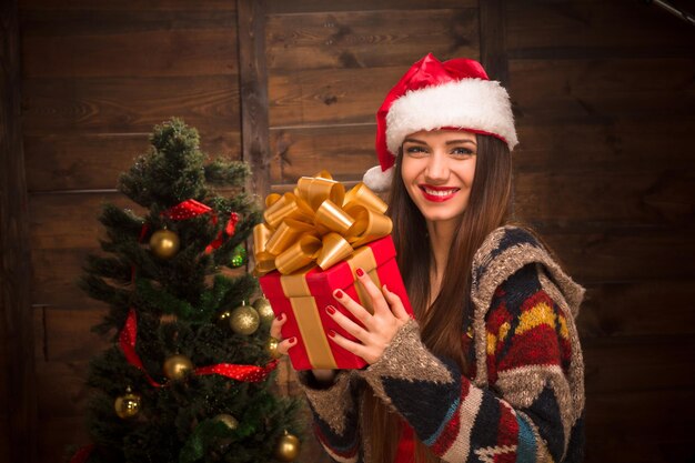 Niña feliz con labios rojos sosteniendo un regalo cerca de Año Nuevo y árbol de Navidad. Hermosa dama con sombrero de Santa mirando a la cámara.