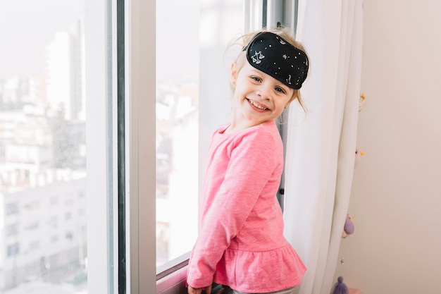 Foto gratuita niña feliz con máscara de ojo para dormir de pie cerca de la ventana de cristal