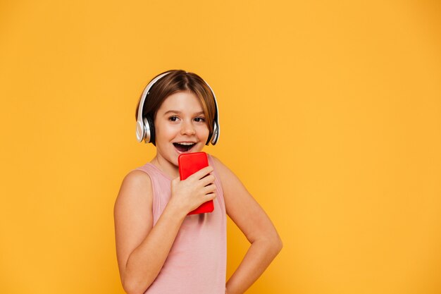 Niña feliz mirando a un lado mientras usa auriculares y cantando en smartphone aislado