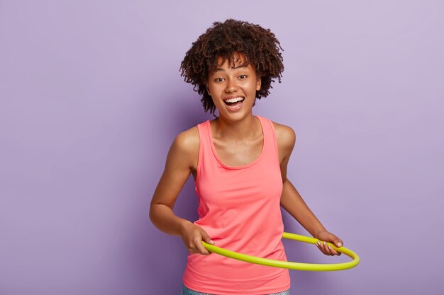 Niña feliz con peinado rizado, gira el hula hoop para adelgazar, tiene ejercicios en el gimnasio, sonríe positivamente, usa chaleco rosa