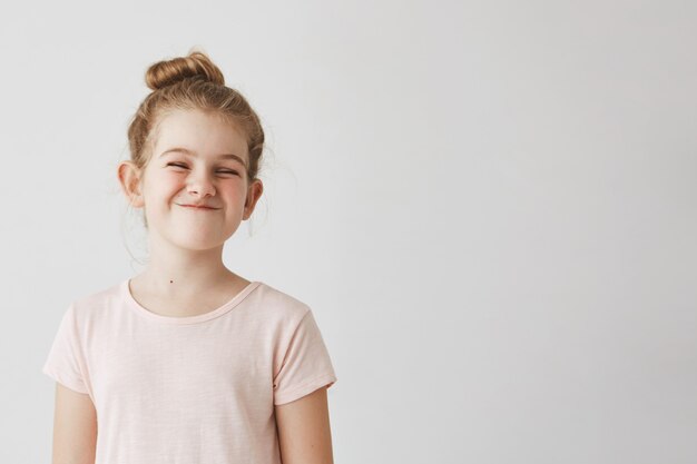 Niña feliz con el pelo largo y rubio en peinado bollo divertido sonriendo con los ojos perdidos, haciendo caras tontas en sesión de fotos de la escuela.