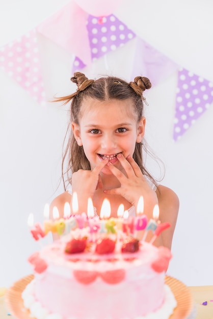 Niña feliz poniendo sus dedos en la boca de pie detrás de pastel de cumpleaños