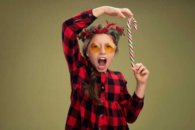 Niña feliz y positiva con corona de navidad en vestido comprobado sosteniendo bastón de caramelo sonriendo alegremente