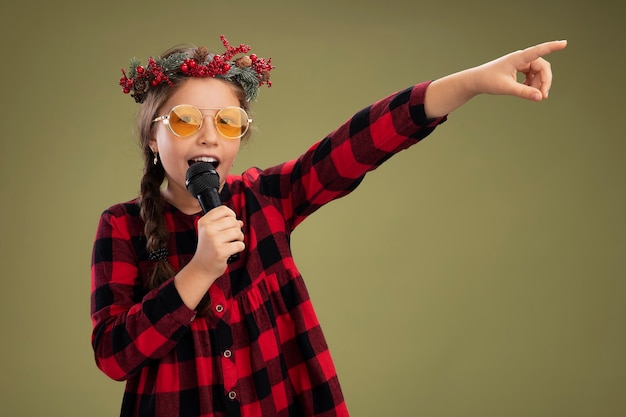 Niña feliz y positiva con corona de navidad en vestido comprobado sosteniendo micrófono gritando apuntando con el dedo índice hacia el lado de pie sobre fondo verde