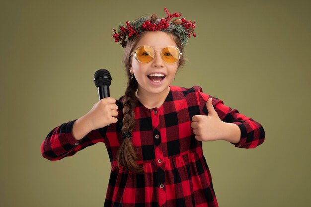 Foto gratuita niña feliz y positiva con corona de navidad en vestido comprobado sosteniendo el micrófono sonriendo alegremente mostrando los pulgares para arriba sobre fondo verde