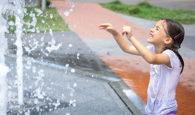 La niña feliz entre las salpicaduras de agua de la fuente de la ciudad se divierte y se escapa del calor.