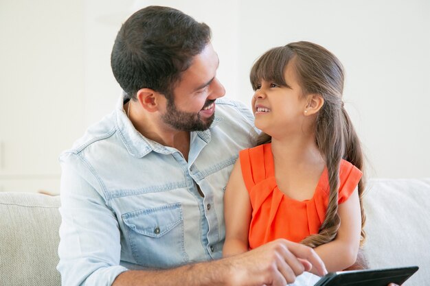 Niña feliz sentada en el regazo de su padre y riendo. Padre disfrutando del tiempo con su hija mientras sostiene la tableta.