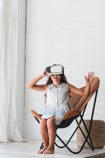 Niña feliz sentada en una silla con gafas de realidad virtual en casa