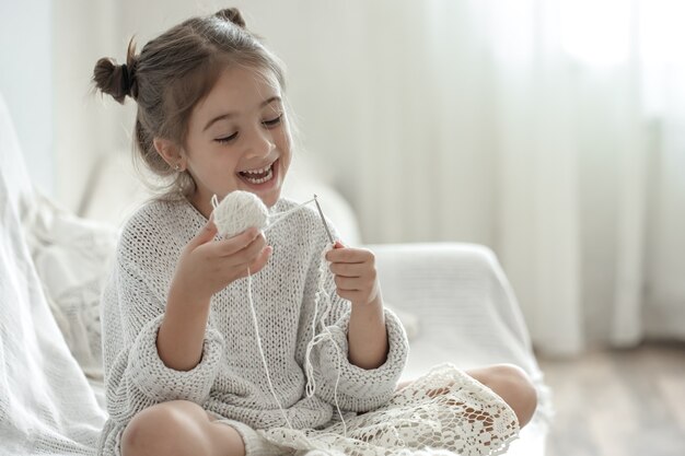 Niña feliz sentada en el sofá y aprendiendo a tejer, concepto de ocio en casa.