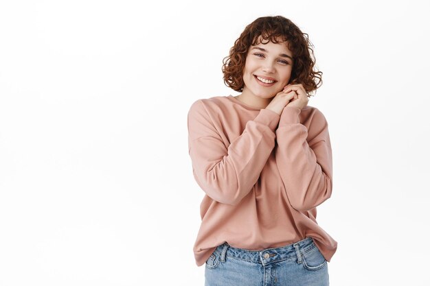 Niña feliz y sincera con una hermosa sonrisa, inclina la cabeza sobre las manos y mira la cámara con una cara soñadora complacida, pensamientos románticos, de pie sobre un fondo blanco. copia espacio