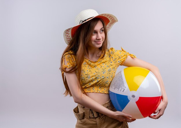 Niña feliz con sombrero sosteniendo una pelota de playa mirando hacia el lado derecho en un espacio en blanco aislado con espacio de copia