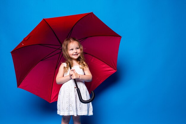 Niña feliz con sombrilla roja posando en la pared azul.