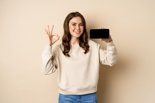 Niña feliz sonriente que muestra la aplicación de pantalla del teléfono inteligente, la interfaz móvil, el signo de OK, de pie sobre un fondo beige.