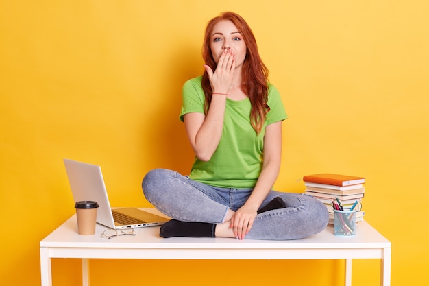 Niña feliz sorprendida, no puedo creer en un triunfo o éxito inesperado, cubre la boca con la palma, usa camiseta verde y jeans, posa contra la pared amarilla del estudio