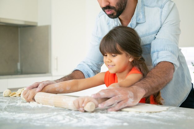 Niña feliz y su papá rodando masa sobre la mesa de la cocina con harina desordenada.