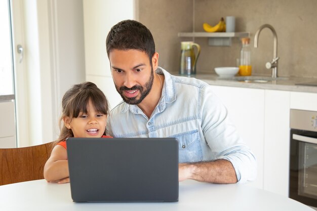 Niña feliz y su papá usando laptop, sentados a la mesa, viendo películas, mirando la pantalla.