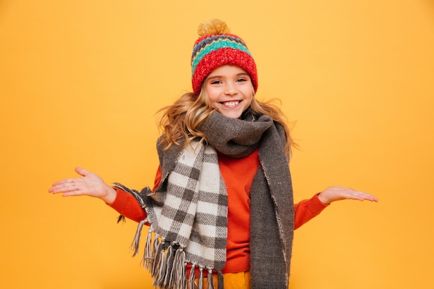 Niña feliz en suéter, bufanda y sombrero se encoge de hombros mientras mira a la cámara