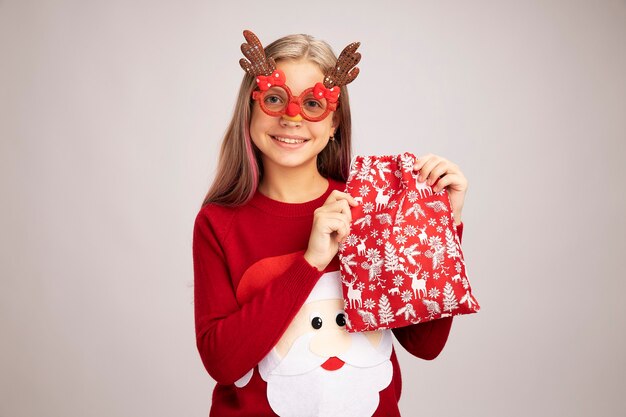 Niña feliz en suéter de Navidad con gafas de fiesta divertidas sosteniendo santa bolsa roja con regalos mirando a la cámara sonriendo alegremente de pie sobre fondo blanco.